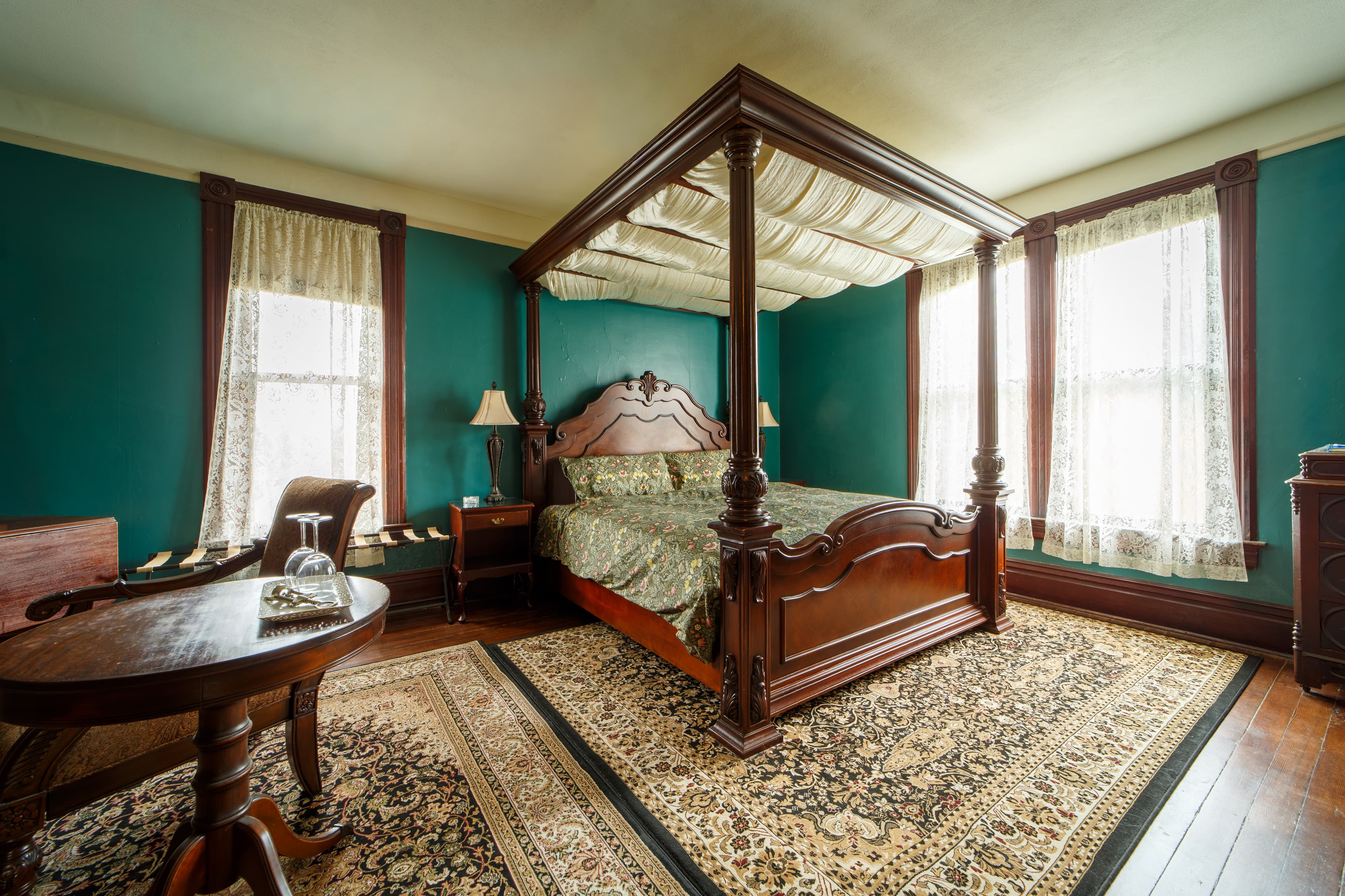 A luxurious guest room featuring a romantic four-poster canopy bed, green wingback armchairs, and a vintage-style sitting area with a small lace-covered table.