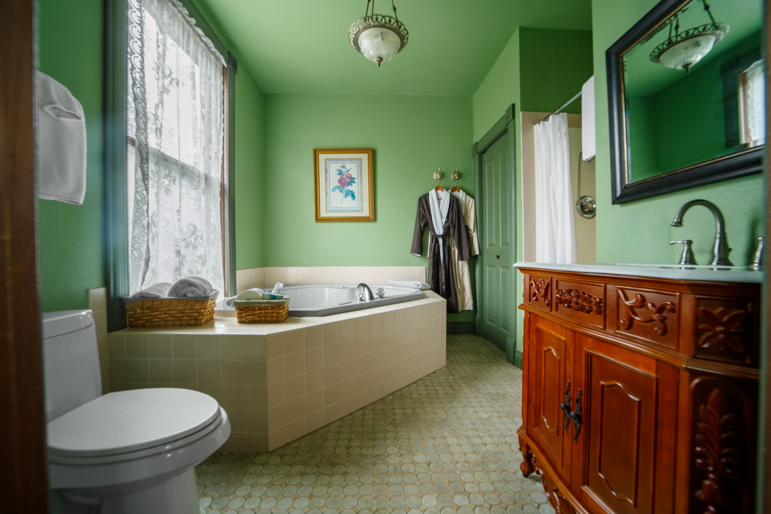 A spacious, green-walled private bathroom featuring a large corner jetted tub, a dark wood vanity with a marble top, and plush bathrobes hanging near a separate shower.