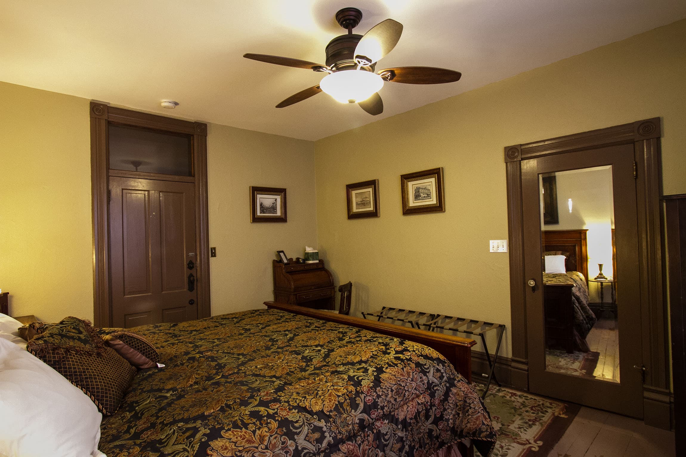A bedroom featuring a dark wood-framed bed with a patterned gold and blue bedspread, small framed artwork on the walls, and a large mirror reflecting the bed.