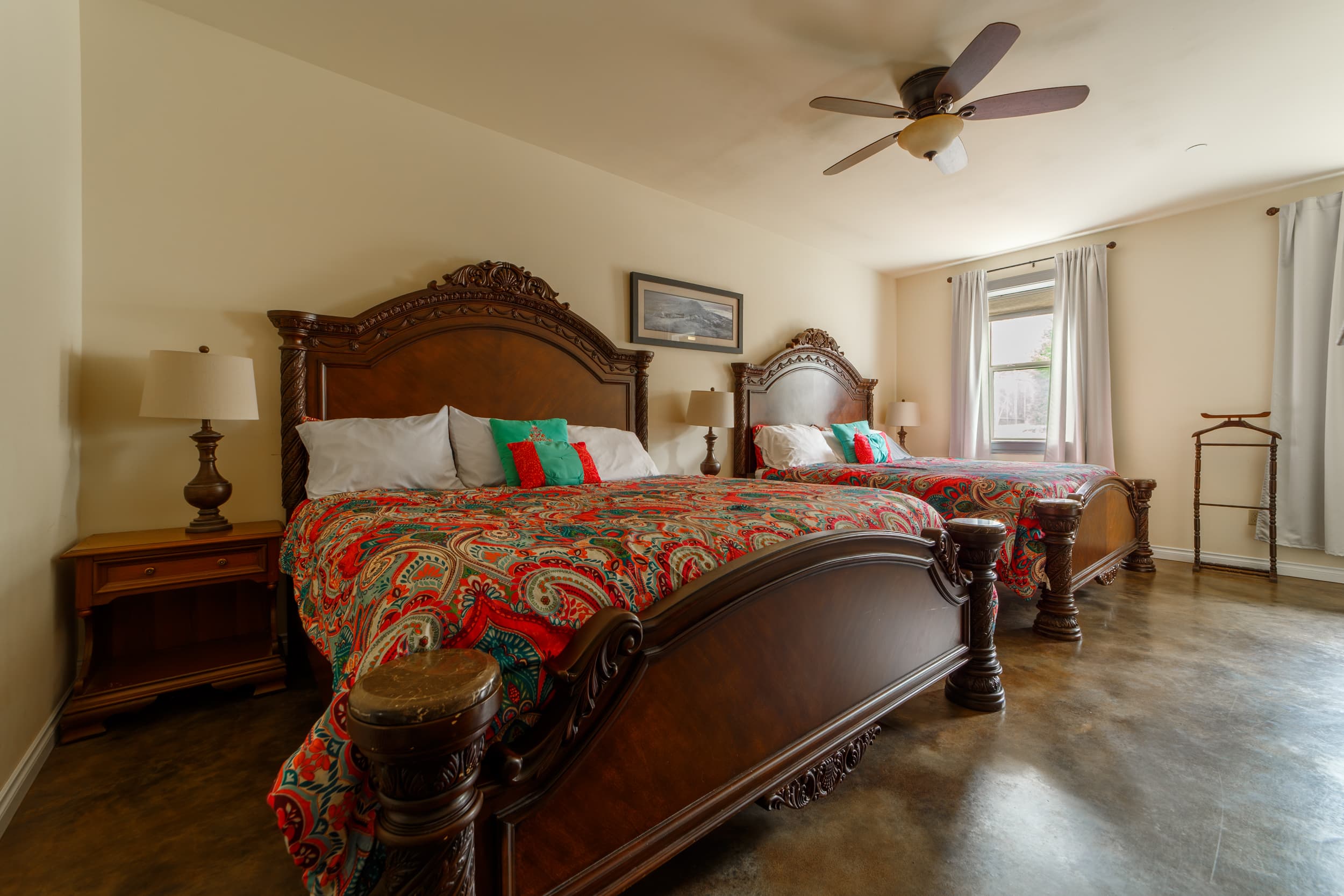 A bright guest bedroom featuring two beds with vibrant pink and teal paisley bedspreads, a wooden dresser, and a vintage vanity desk.