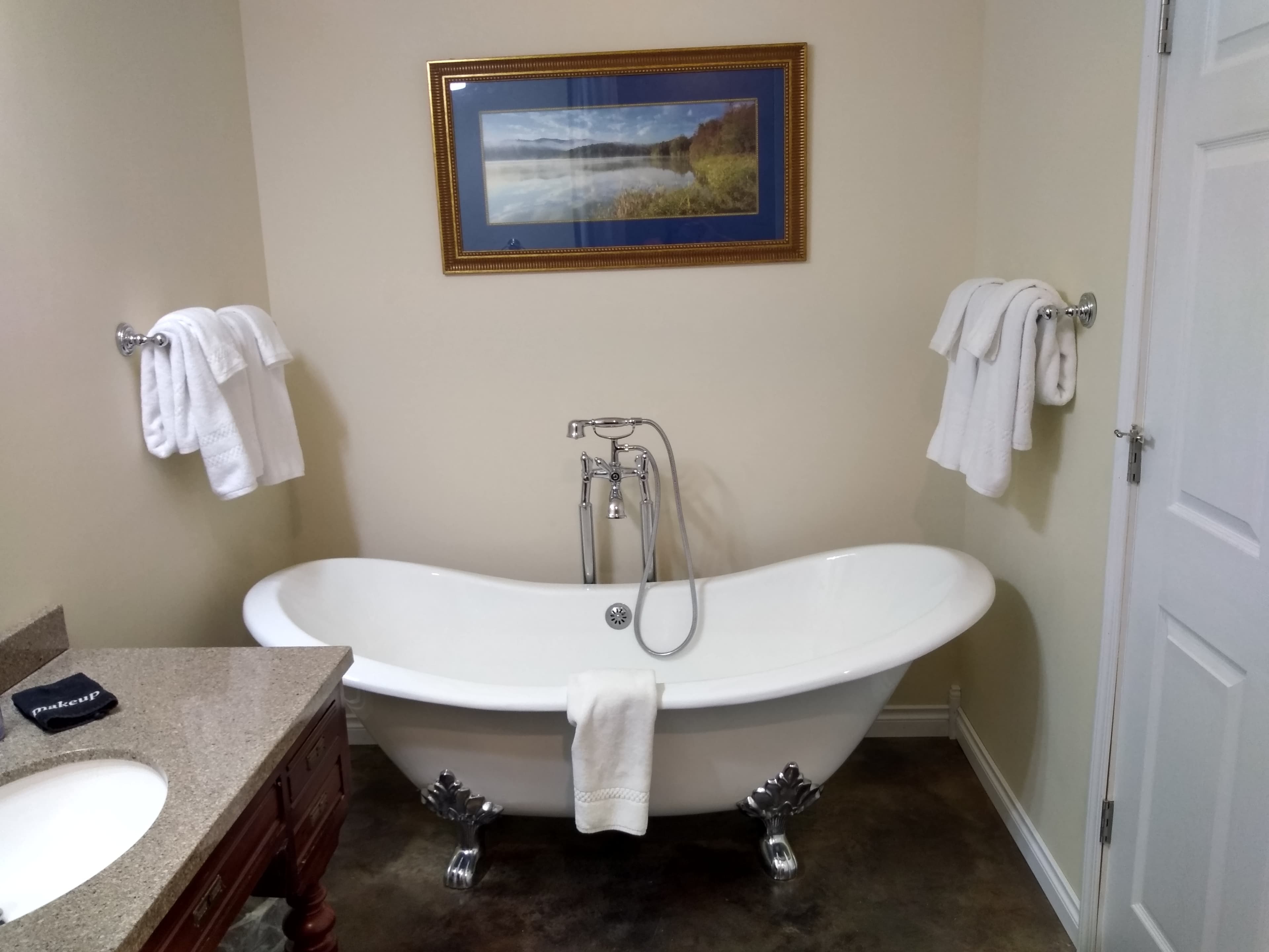 A white, vintage-style clawfoot soaking tub with a silver hand-held shower attachment, situated in a modern bathroom with beige walls, a dark vanity, and white towels.
