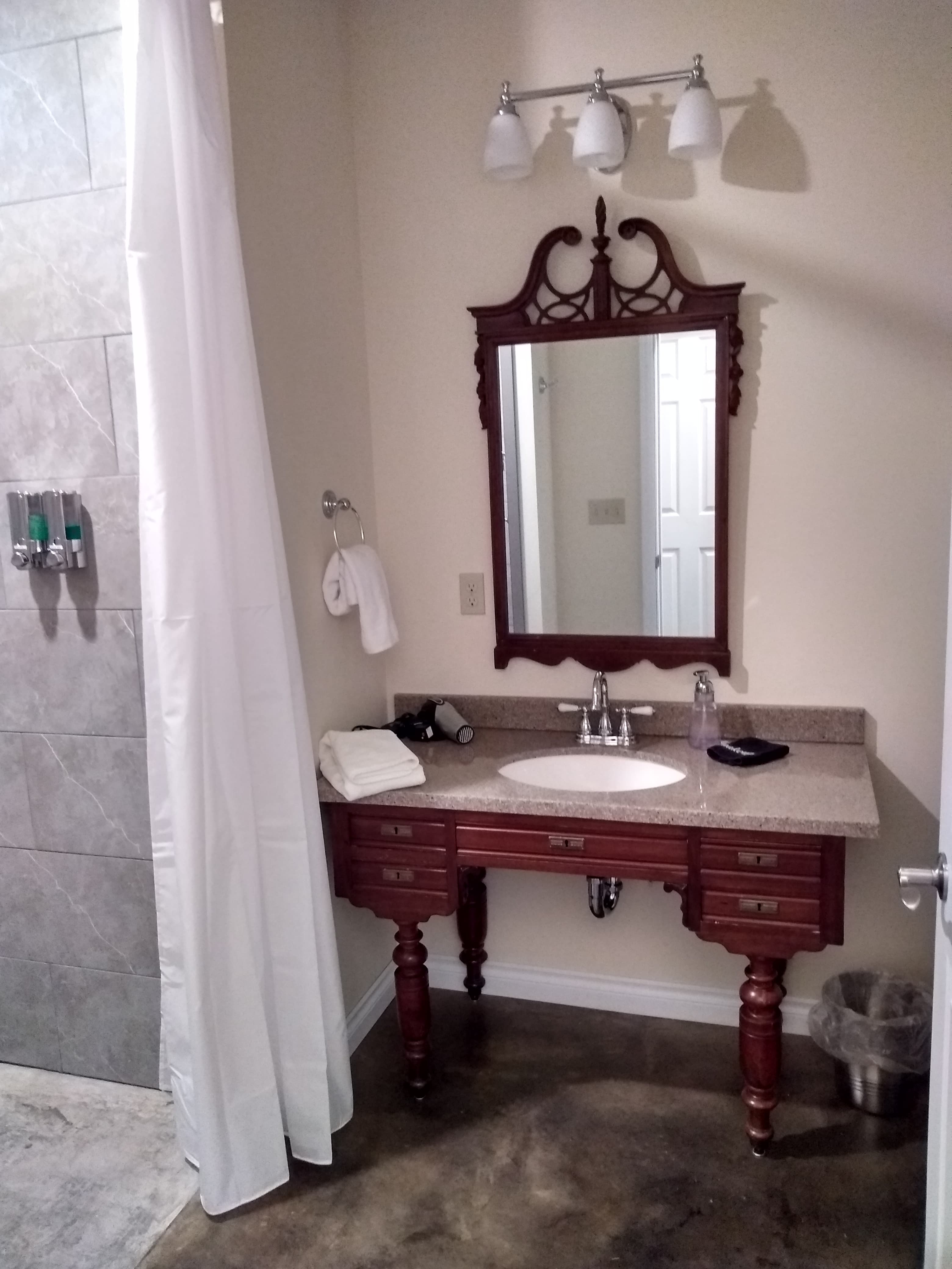 A classic wood vanity with an ornate, dark wood-framed mirror and three frosted glass lights, situated next to a stone-tiled walk-in shower with a white curtain.