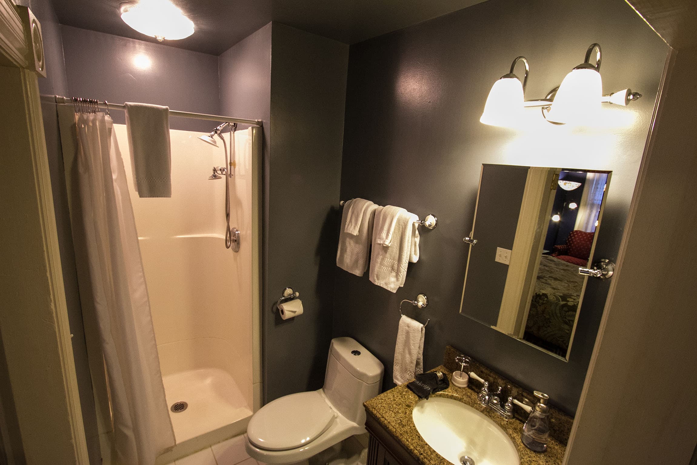 A high-angle view of a modern private bathroom featuring a white walk-in shower with a curtain, a toilet, and a vanity with a granite countertop and lighted mirror.