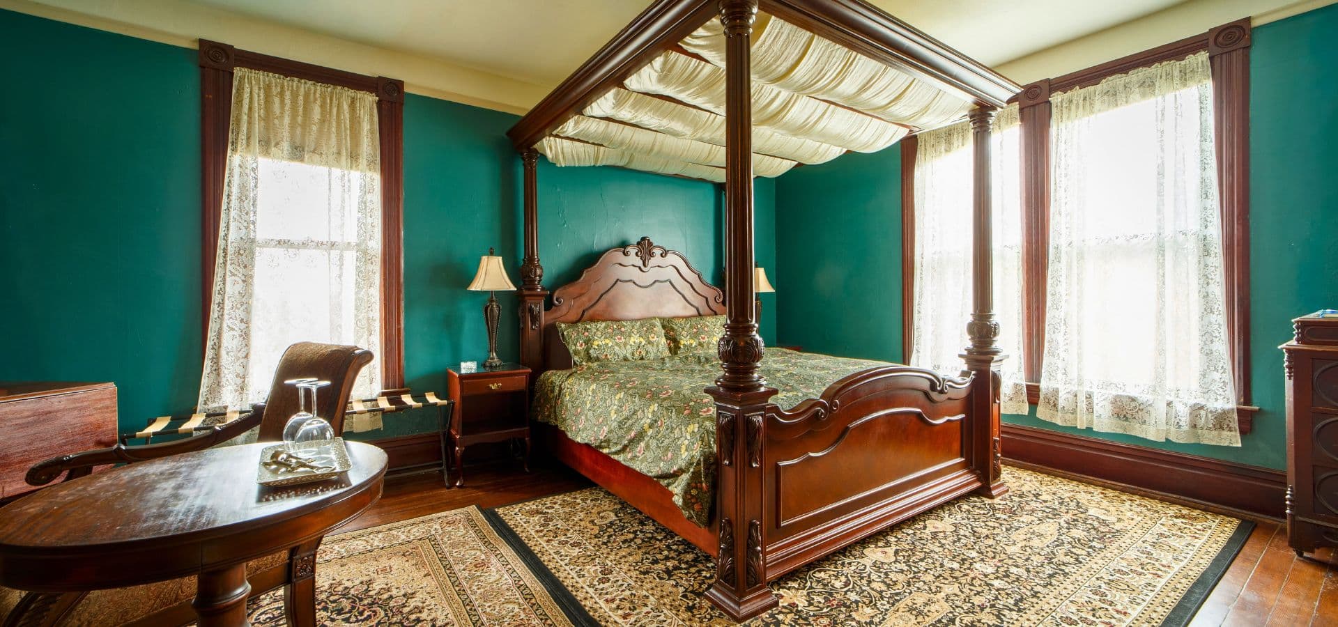 A luxurious bedroom featuring a four-poster bed, dark wood furniture, and teal walls with large windows.