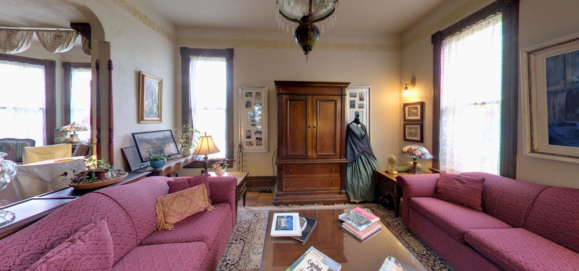 A cozy, vintage living room featuring burgundy sofas, a wooden armoire, and large windows with light streaming in.